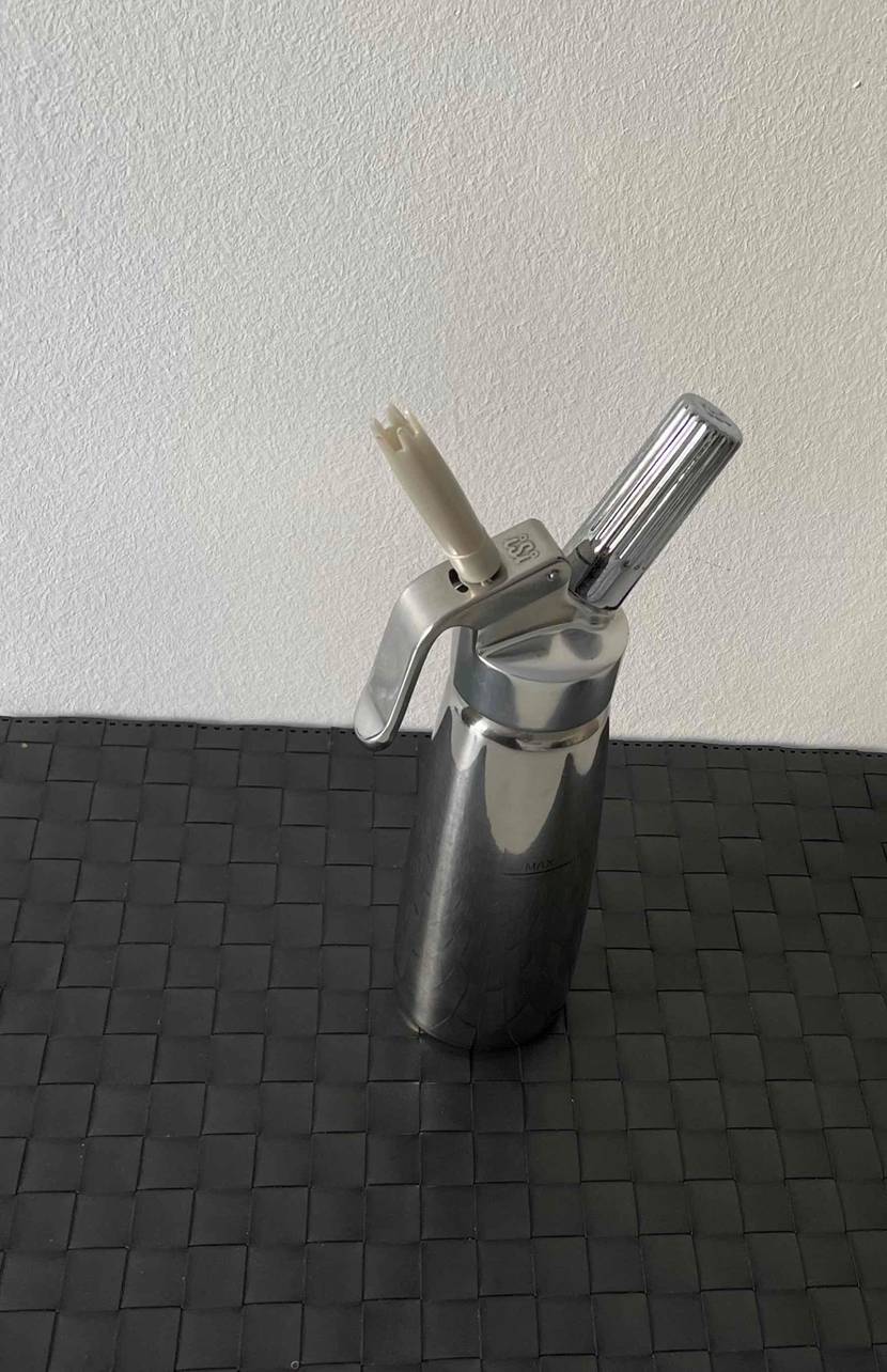 Ein getesteter Sahnespender von schräg oben auf einer dunklen Fläche stehend.