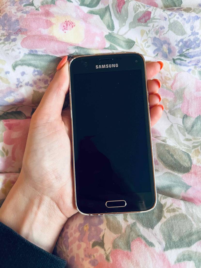 Ein ausgeschaltetes, getestetes kleines Smartphones von Samsung in der Hand auf einer geblümten Stoffunterlage.