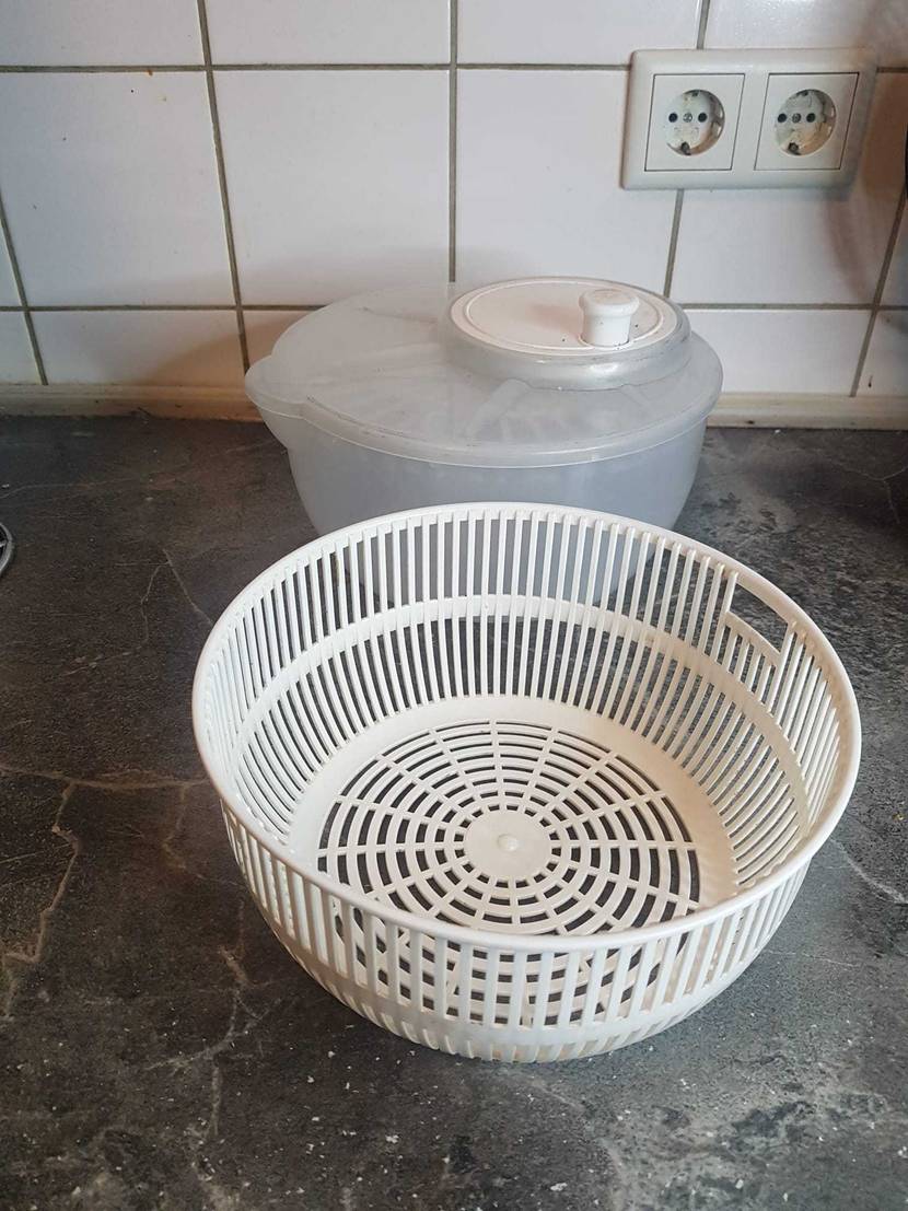 Salatschleuder im Test: ein weißes Innensieb steht vor einem transparenten, runden Kunststoffbehälter inklusive Deckel mit weißer Kurbel, auf einer grauen Marmorplatte vor einem weißen Fliesenspiegel mit zwei Steckdosen.