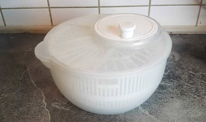 Eine getestete Kunststoff-Salatschleuder im transparenten Design steht inklusive weißem Innensieb und Deckel mit weißer Kurbel auf einer grauen Marmorplatte.