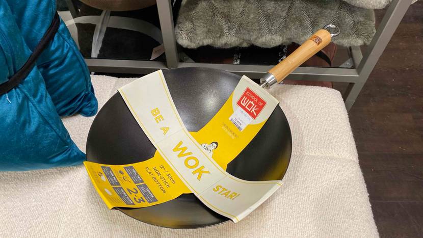 Ansicht von oben auf einen grauen Wok im Test, mit Holzgriff und Originalverpackung auf hellem Hocker in einem Geschäft.