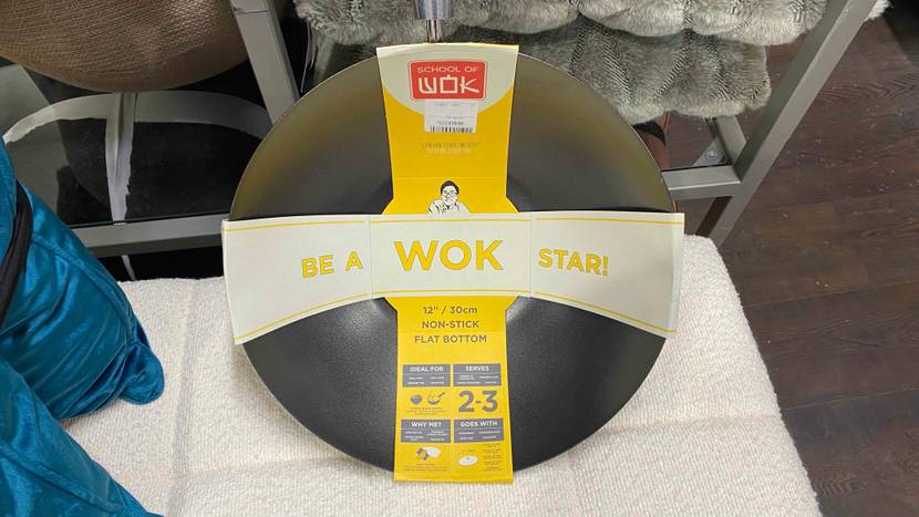Wok im Test der Marke Wok of School befindet sich in einem Geschäft auf einem mit beigefarbenen Stoff bezogenen Hocker.