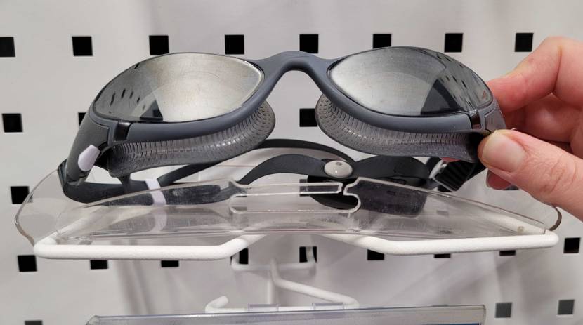 Hand greift nach einer vor weißem Hintergrund ausgestellten grauen Schwimmbrille im Test, die über entspiegelte getönte Kunststoffgläser verfügt.