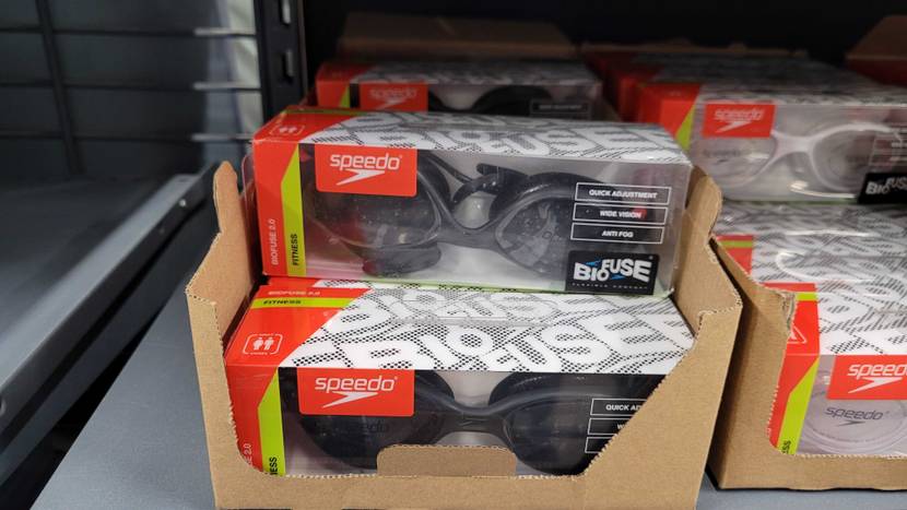 Mehrere Kunststoffverpackungen mit Speedo Biofuse Schwimmbrillen im Test liegen gestapelt in braunen Kartons im Verkaufsregal.