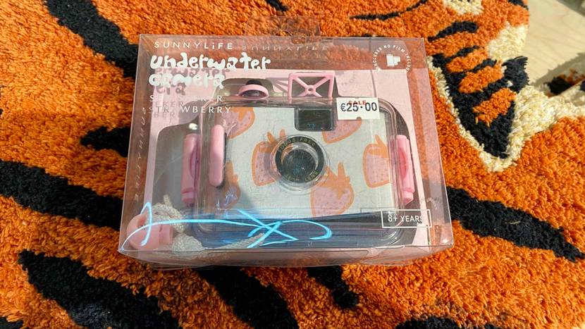 Eine Unterwasserkamera im Test, mit rosa Erdbeeren bedruckt, befindet in ihrer durchsichtigen Kunststoffverpackung auf einem orange-schwarzen Teppich.