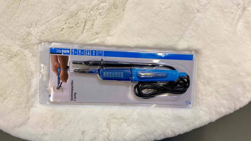 Ein digitaler, blauer Unitec Spannungsprüfer im Test befindet sich in seiner Originalverpackung auf einem weißen Langhaarteppich.