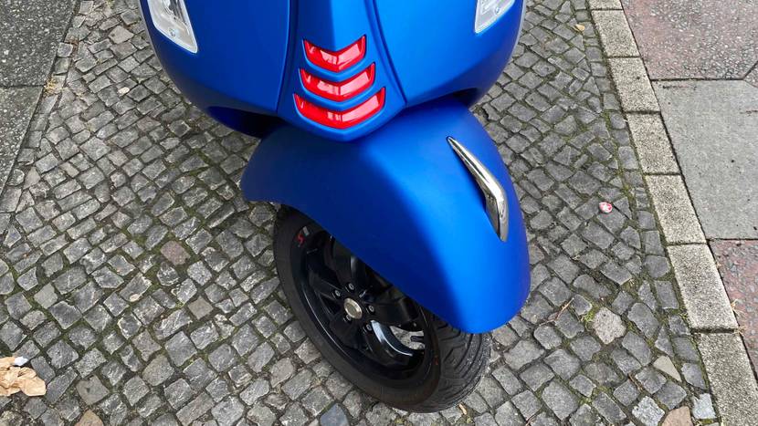 Vorderreifen und Blinker eines auf Kopfsteinpflaster stehenden blauen Vespa Motorrollers im Test.