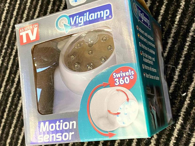 Nahaufnahme eines 360-Grad-Vigilamp Bewegungsmelders im Test, der sich innerhalb seiner Originalverpackung auf einer gestreiften Türmatte befindet.
