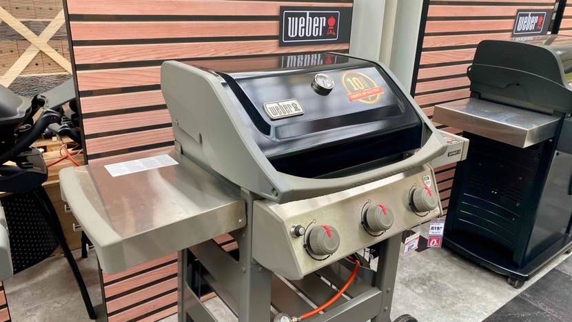 Ein hellgrauer Weber Stand-Gasgrill im Test befindet sich mit abgedeckter Grillfläche neben einem weiteren Modell in einem Geschäft.