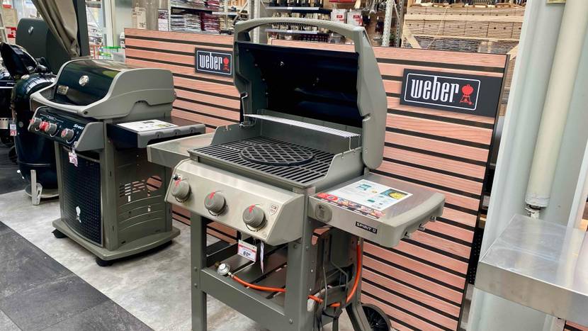 Ein Weber Stand-Gasgrill im Test steht mit geöffnetem Grillbereich, mit anderen Grill-Modellen, in einem Verkaufsbereich.