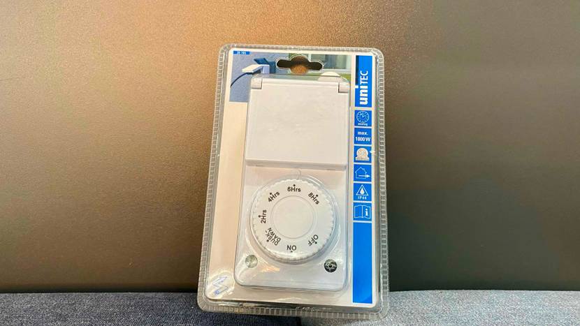 Eine transparent verpackte Zeitschaltuhr lehnt an einer grauen Wand.